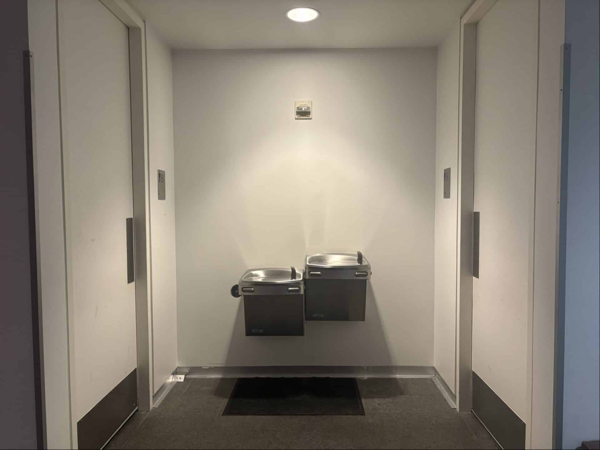 Two silver drinking fountains are mounted on a white wall between two closed restroom doors under a ceiling light in a hallway.