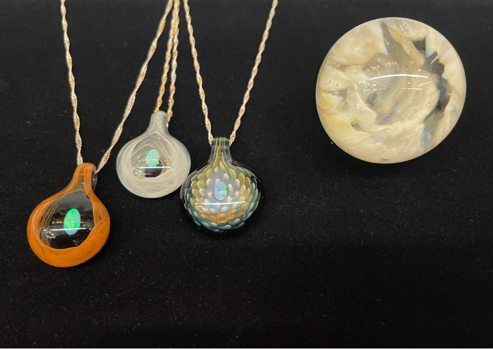 Three glass pendants with iridescent centers on twisted cords and a round glass paperweight with cloudy swirls are displayed on a black surface.