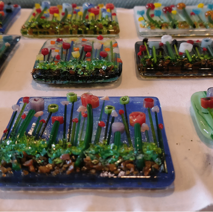 Colorful glass tiles decorated with raised, three-dimensional designs of flowers and grass, arranged on a flat surface. Each tile features a variety of bright flowers and green stems.