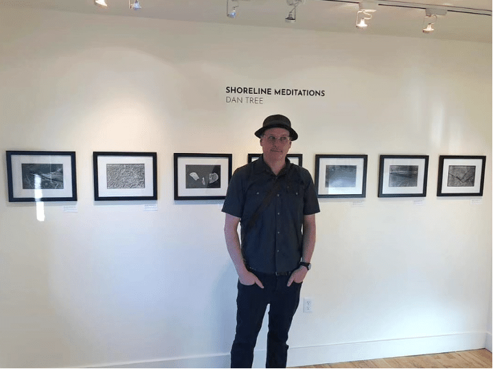 A person wearing a hat and glasses stands in front of a gallery wall displaying six framed black-and-white artworks under the exhibit title Shoreline Meditations by Dan Tree.