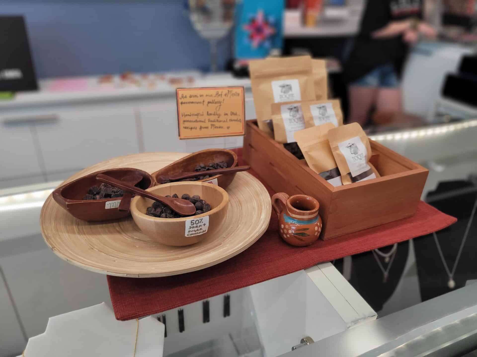 A display of chocolate samples in wooden bowls with labels, a small pitcher, and packaged chocolates on a red mat atop a glass counter in a shop.