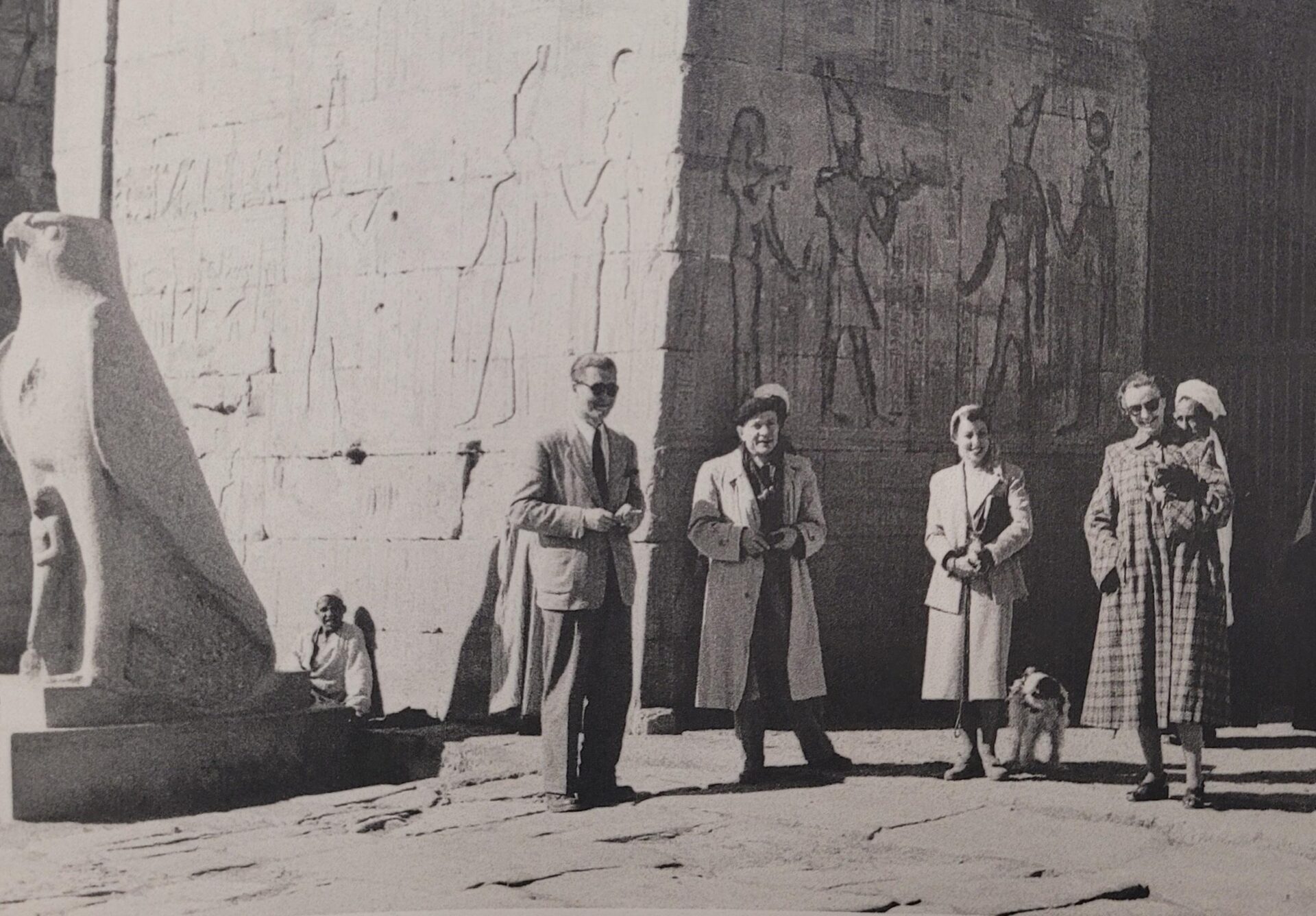 Five people and a small dog stand in front of an ancient stone wall with Egyptian carvings. A large statue of a falcon is visible to the left. The image is in black and white.