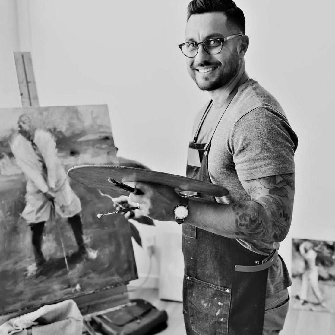 A smiling artist wearing glasses and an apron holds a palette while painting a golfer on a canvas in a well-lit studio.